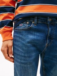 Pantalones vaqueros de denim azul con un corte recto, que presentan costuras visibles y una etiqueta de logo. Combinados con una camisa de manga larga de rayas en azul marino y naranja.