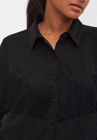 Chemise noire en tissu lisse avec un design frangé sur la poitrine, dotée d'un col classique et d'une fermeture à boutons.