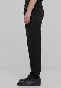 Črne jeans hlače s ravnim krojem, ki vsebujejo subtilne poškodbe in vidne šive. Ustrezajo z črnimi športnimi copati.