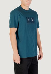 T-shirt en coton couleur sarcelle avec un graphique portant le texte "AX" et "Armani Exchange". Design à manches courtes et col rond.