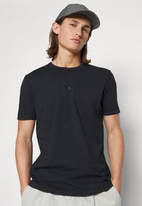 T-shirt preta de manga curta com gola redonda e tecido texturizado. Apresenta um pequeno logótipo preto no peito. Combinada com um boné cinza estampado.