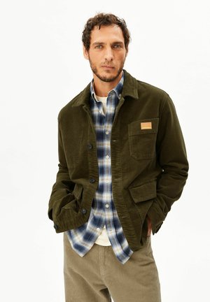 Mann trägt eine olivgrüne Cordjacke über einem blau-beigen karierten Hemd mit beigen Cordhosen, steht mit den Händen in den Taschen.