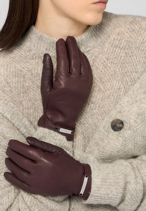 Kobieta w ciemnobrązowych skórzanych rękawiczkach i beżowym dzianinowym swetrze z dużym guzikiem, z rękami skrzyżowanymi przy klatce piersiowej.