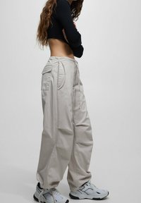 Person med långt hår, iförd baggy beige byxor, vita sneakers och en svart kortärmad topp, stående på sidan mot en enkel bakgrund.