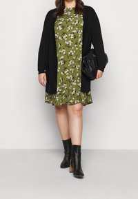 Cardigan nero su un vestito floreale verde con fiori bianchi e gialli. Stivali alla caviglia neri con tacco a blocco. Borsa nera.