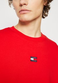 Piros pulóver kerek nyakkivágással, puha anyagból készült. A mellkasán egy kis fekete címke található "TOMMY JEANS" logóval.