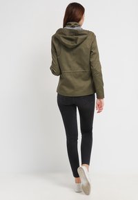 Olivově zelená bunda s kapucí, s hladkou texturou a přiléhavým střihem. V kombinaci s černými skinny džíny a bílými teniskami.