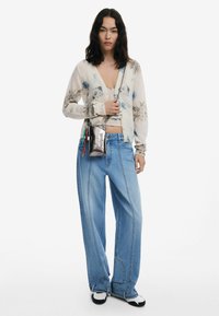 Cardigan surdimensionné à motifs floraux en crème, avec des fleurs bleues. Jean large bleu clair, associé à des chaussures noires et blanches et un sac bandoulière en argent.