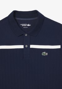 Polo de Lacoste en azul marino, con textura acanalada, tapeta de dos botones y una franja horizontal blanca en el pecho con acento de logo.