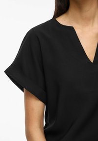 Top nero a scollo a V con maniche corte arrotolate, realizzato in un tessuto liscio. Presenta una vestibilità comoda e un design minimalista. Nessun motivo o accentuazione visibile.