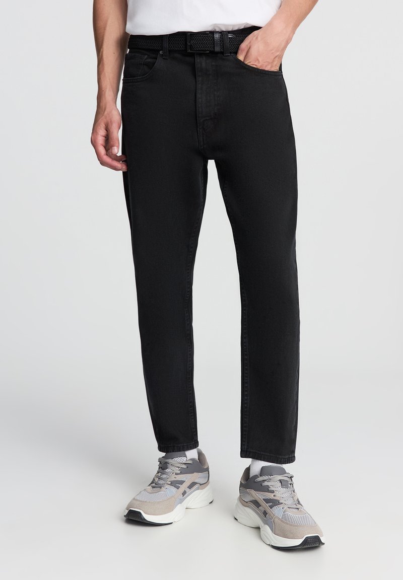 Jeans in denim nero con una vestibilità dritta, design a cinque tasche e una cintura, abbinati a scarpe da ginnastica grigie e bianche.