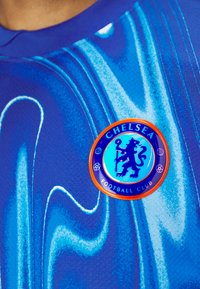 Blå tröja med grafisk design som visar svängningar i olika nyanser av blått. Inkluderar en cirkulär logotyp för Chelsea Football Club på bröstet.