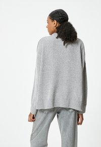 Grauer Strickpullover mit lockerer Passform, geripptem Saum und Rückendetail, kombiniert mit hellgrauen Jeans. Weiche Textur und entspannter Schnitt.