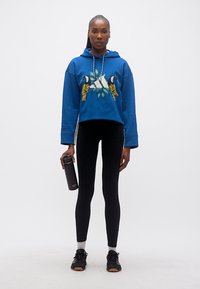 Γυναίκα που στέκεται φορώντας μπλε φούτερ με κουκούλα και γραφικό πουλιού, μαύρο κολάν, μαύρα αθλητικά παπούτσια, κρατώντας μαύρο παγούρι νερού.