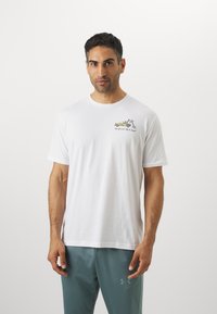 Mand iført hvid T-shirt med grafisk design af en gul bugserbil og tekst, parret med teal Under Armour bukser, står op ad en ensfarvet hvid baggrund.