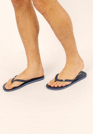 Des pieds portant des tongs bleu marine avec des rayures blanches, montrant des jambes poilues sur un fond clair.