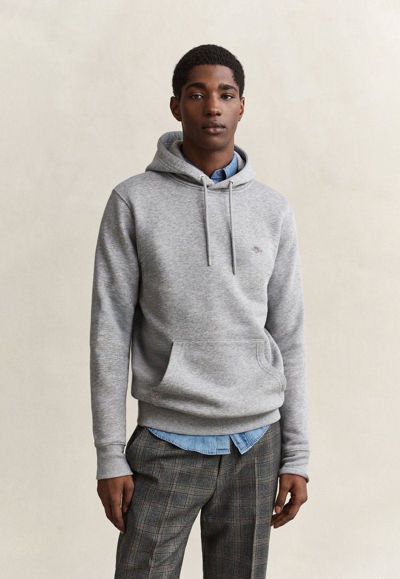 Jeune homme portant un sweat à capuche gris par-dessus une chemise bleue et un pantalon à carreaux, debout devant un fond uni clair.
