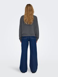 Kvinde med langt bølget hår står med ryggen mod og bærer en mørkegrå sweatshirt og blå jeans med vide ben foran en ensfarvet baggrund.