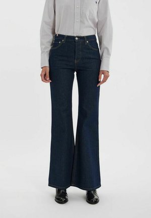 Jean flare taille haute en denim foncé, avec des coutures contrastantes, un design à cinq poches et une texture lisse. Accompagné d'une chemise rayée.