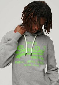 Felpa grigia con stampa grafica verde, cappuccio con cordino e polsini a costine. Include una tasca anteriore e un'etichetta con il logo sulla manica.