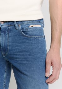 Blauwe spijkerbroek met een slanke pasvorm, met een voorzak waarin een smartphone is gestoken. De stof heeft een gladde textuur met subtiele vervaging.