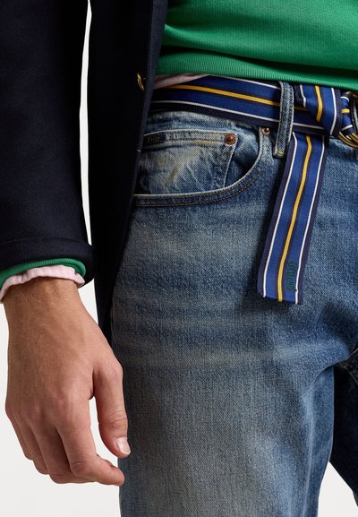 Jeans in denim di un blu sbiadito, con una cintura a righe blu e gialla. Sovrapposti con una camicia verde e un blazer blu scuro.