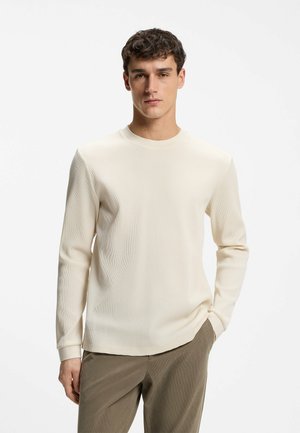 Junger Mann mit kurzem, dunklem Haar, der einen cremefarbenen, strukturierten Langarmpullover und braune Hose trägt, mit einer Hand in der Tasche stehend.