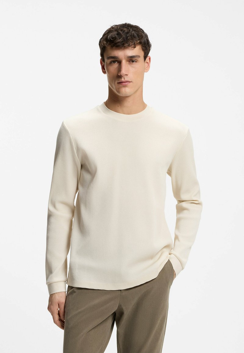 Jeune homme aux cheveux courts et foncés, portant un pull texturé crème à manches longues et un pantalon marron, debout avec une main dans la poche.