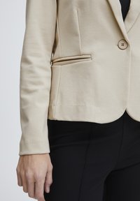 Béžový blazer z hladkého materiálu, s jedním knoflíkem, boční kapsou a přiléhavým střihem. Nosený přes černé kalhoty, s upraveným vzhledem.