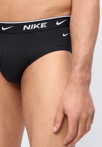 Κοντινό πλάνο ενός ατόμου που φορά μαύρα εσώρουχα Nike με λευκό λογότυπο στη μέση και στο πόδι, εμφανίζοντας τον κορμό και το δεξί χέρι.