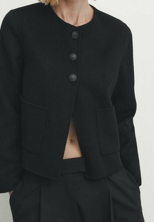 Black long-sleeve buttoned jacket with two front pockets worn over black pleated pants, showing a small midriff section.
