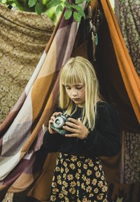 Kind met lang blond haar houdt een zwarte camera vast. Ze draagt een zwarte top met textuur en een rok met bloemenpatroon. De achtergrond heeft kleurige gordijnen.