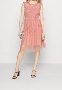 Vestido con encaje en suave rosa, con un corsé ajustado y una falda fluida y transparente. Combinado con tacones negros de tiras y un clutch plateado.