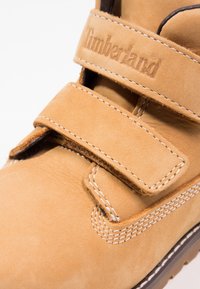 Botte en cuir tan avec une sangle en velcro, couture blanche et semelle robuste. Présente le logo "Timberland" embossé sur la sangle.