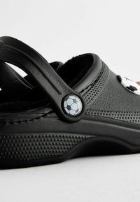 Zueco negro con interior de felpa, superficie texturizada y un botón circular con un diseño de balón de fútbol. Vista lateral en ángulo destacando los detalles.