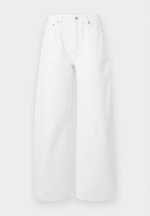 Vrouw draagt een crème geribde cardigan met knopen, wijde witte broek, bruine riem en crème slingback ballerina's, staand met één hand in de zak.