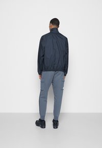 Marine windbreaker met hoge kraag, elastische manchetten en tailleband, gecombineerd met grijze cargo broek met zijzakken en een streepdetaillering.
