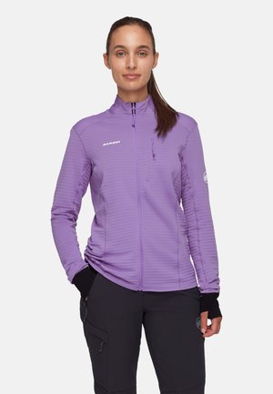 Vrouw met bruin haar in een paardenstaart en een paarse jas met rits en zwarte broek, staand tegen een effen lichte achtergrond.