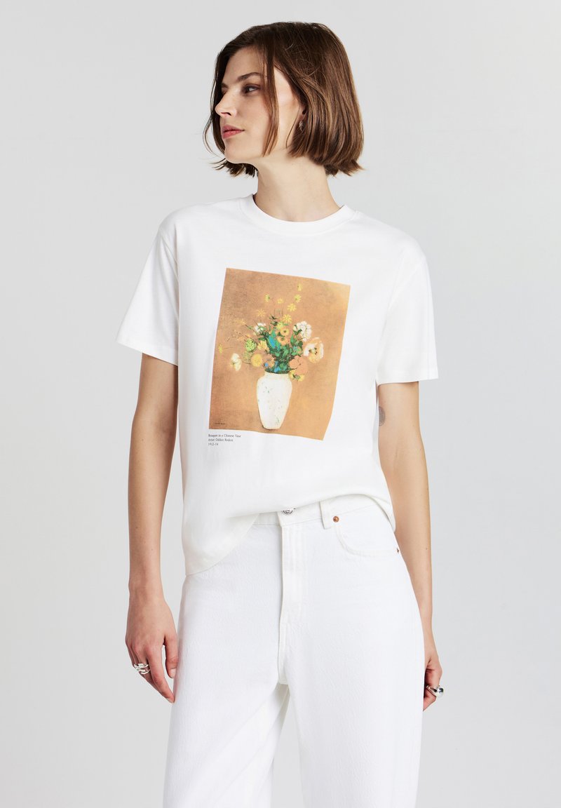 Donna con capelli corti castani, che indossa jeans bianchi e una maglietta bianca con una pittura di un vaso floreale sulla parte anteriore, che guarda a sinistra.