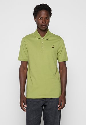 Mand iført lysegrøn kortærmet poloshirt med gul fuglelogo og mørkegrå jeans, stående mod en ensfarvet lys baggrund.