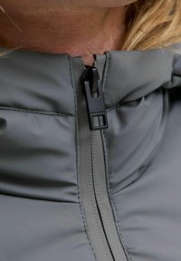 Szara kurtka z zamkiem błyskawicznym, charakteryzująca się gładką, matową teksturą, stójką oraz czarnym uchwytem zamka z detalem marki.