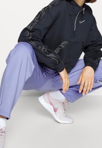 Schwarzer Quarter-Zip-Sweatshirt mit weißen Ziernähten, kombiniert mit lavendelfarbenen Jogginghosen. Hellrosa Sneaker runden den Look ab.
