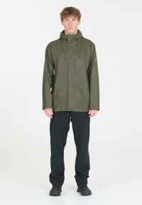 Wasserdichte olivgrüne Jacke mit Kapuze, ausgestattet mit einem Druckknopfverschluss und zwei Vordertaschen, kombiniert mit schwarzen Hosen und dunklem Schuhwerk.