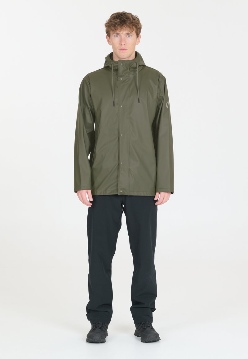 Wasserdichte olivgrüne Jacke mit Kapuze, ausgestattet mit einem Druckknopfverschluss und zwei Vordertaschen, kombiniert mit schwarzen Hosen und dunklem Schuhwerk.