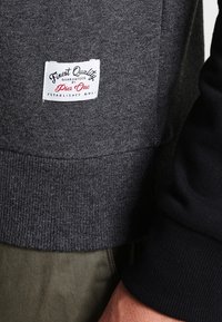 Grå sweatshirt med ribbad textur, med en etikett som säger "Finest Quality by Piec One." Svarta detaljer på ärmarna.