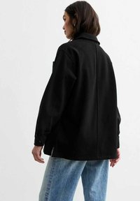 Giacca nera oversize con collo e polsini abbottonati, realizzata in un tessuto morbido e texturizzato. Presenta una cucitura centrale e una vestibilità rilassata.