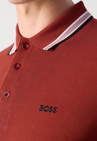Gros plan d'une personne portant un polo couleur rouille avec un col rayé et le logo noir "BOSS" sur la poitrine.