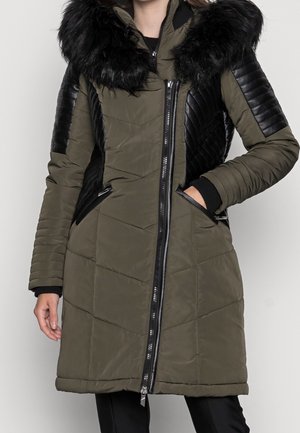 Femme portant un manteau matelassé vert olive avec des panneaux en cuir noir sur les manches et les côtés, un grand col en fausse fourrure noire et une fermeture éclair avant.