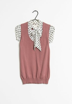 Haut en tricot sans manches, couleur rose poussiéreux, avec un col arrondi ; superposé à un chemisier blanc à pois et avec un nœud fixé au cou.