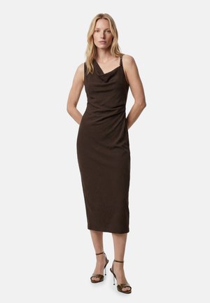 Kobieta o blond włosach, ubrana w ciemnobrązową sukienkę midi bez rękawów oraz oliwkowo-zielone sandały na wysokim obcasie, stojąca z rękami za plecami.